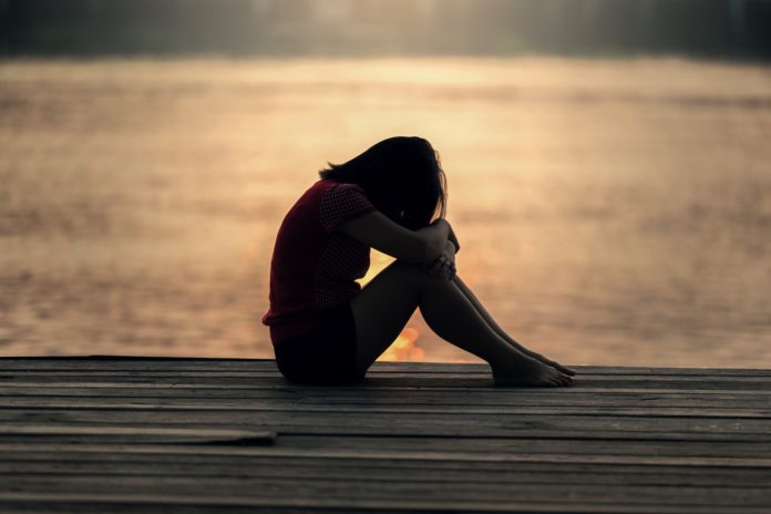 A sad young girl sitting on a pier with her head resting on her knees.