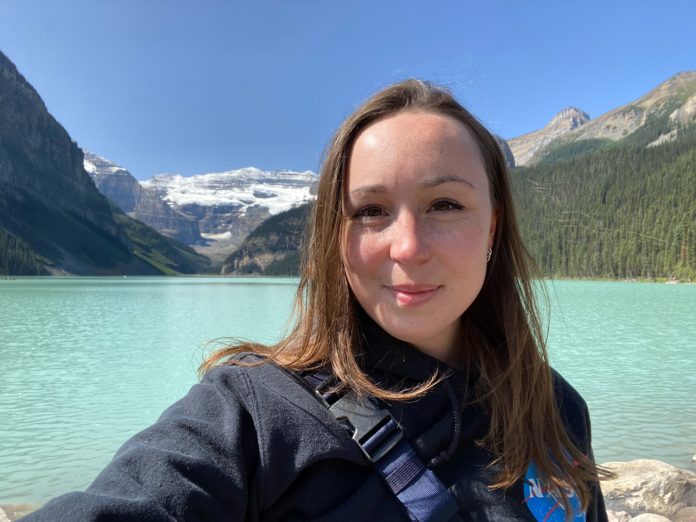 A portrait of Lysa standing in front of a bright blue lake and snow-covered mountains.