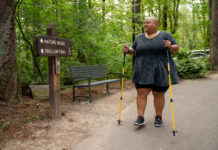 A disabled Black non-binary hiker uses trekking poles and looks at a wooden sign with “Nature House” and “Trillium Trail ♿” marked ahead. The shot is framed with the hiker walking toward the camera and the hiker sports a shaved head, glasses, a gray peplum shirt, black shorts, and black tennis shoes.