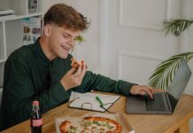 A university students sits in front of a laptop, eating a pizza and drinking a Coca-Cola.