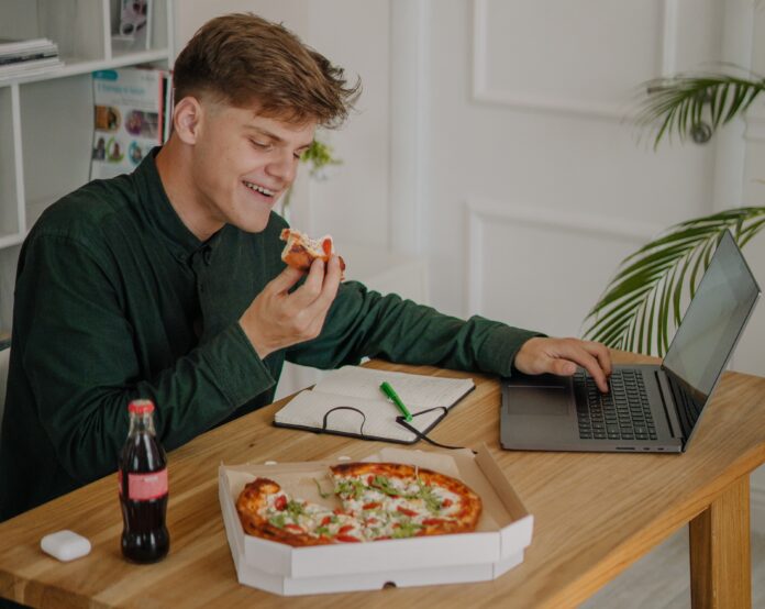 A university students sits in front of a laptop, eating a pizza and drinking a Coca-Cola.