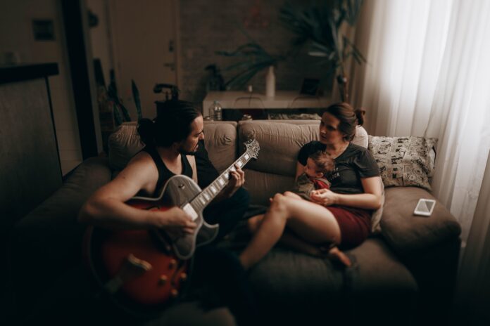 A baby, held by an adult, listens to another adult playing a song on a guitar.