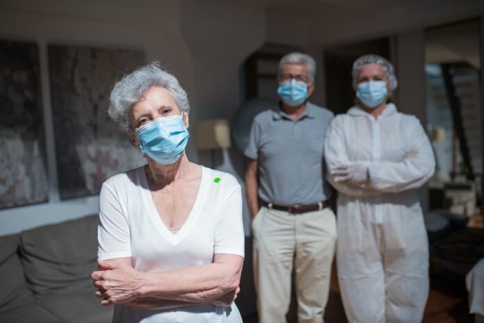 An elderly woman wearing a medical mask looks into the camera. Two other elderly people in medical masks stand behind her.