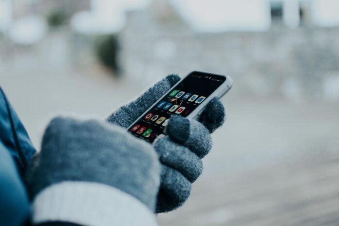 A close-up photo of someone's hands holding a cell phone. The hands are wearing blue gloves. Various social media app icons can be seen on the phone screen.
